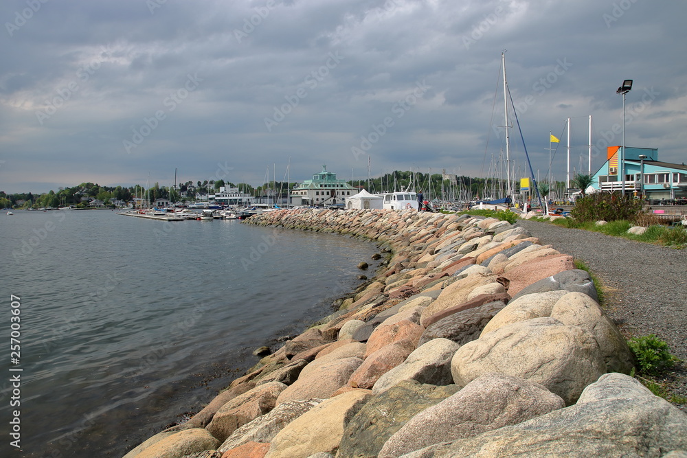 Sea bay, stony shore in Oslo, Norway, boats, yachts, buildings in background, cloudy sky