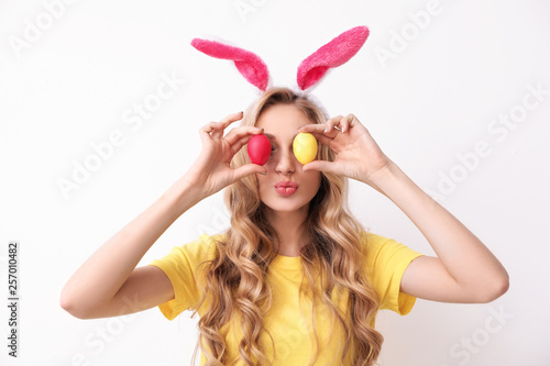 Beautiful young woman in bunny ears headband holding Easter eggs near eyes on light background