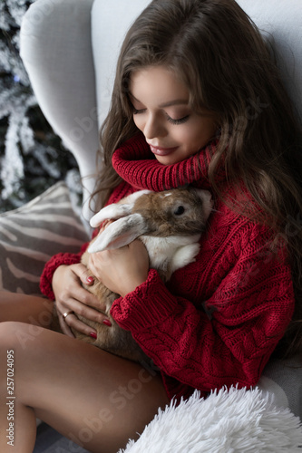 Beautiful leggy young girl  wearing red sweater and wool socks sits at the window and gently holds a rabbit in her hands in christmas interior. Commercial and advertising design