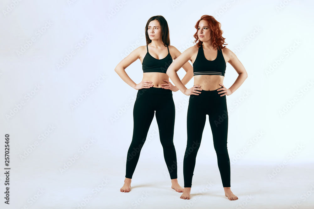 Portrait of two happy young women with different hair color, standing in sportswear together, looking at aside isolated over gray background. Sport, Fitness and Healthy Lifestyle. Portrait of two happy young women with different hair color, standing in sportswear together, looking at aside isolated over gray background. Sport, Fitness and Healthy Lifestyle.