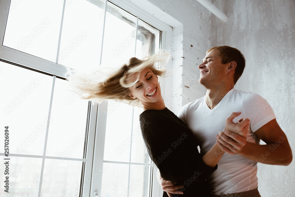 A pair of lovers, hug, kiss, laugh and have fun. The girl in a long black sweater and white high golf. A guy in a white T-shirt and trousers. Light gray interior, large window, brown stylish chair. A pair of lovers, hug, kiss, laugh and have fun. The girl in a long black sweater and white high golf. A guy in a white T-shirt and trousers. Light gray interior, large window, brown stylish chair.