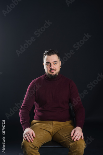 a man with a beard in casual clothes poses on an isolated dark background, in a burgundy sweater in the studio