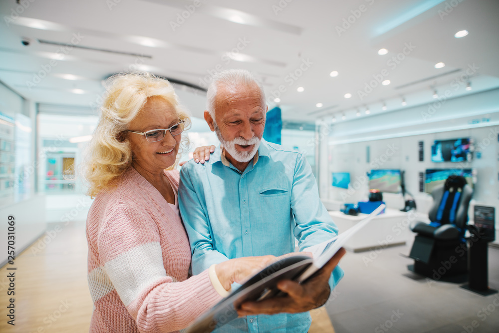 Fototapeta premium Happy Caucasian senior couple looking at brochure and choosing what to buy while standing in tech store.