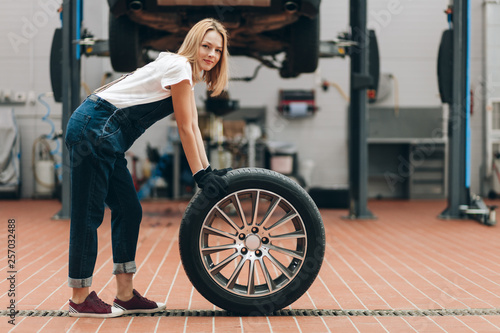 attractive blond girl with a wheel looking at the camera in the repair shop. full length photo. lifestyle. girl repairing a car