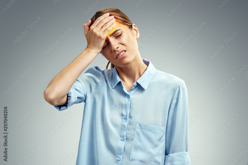 Tired girl suffering from painful headache and stress. Photo of american girl in blue shirt holding hand on forehead on gray background. Medical concept Tired girl suffering from painful headache and stress. Photo of american girl in blue shirt holding hand on forehead on gray background. Medical concept