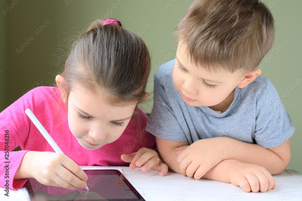 White caucasian toddler boy and beautiful girl drawing together on tablet screen. Portrait of a happiness little brother and sister. Children play a computer game on the tablet White caucasian toddler boy and beautiful girl drawing together on tablet screen. Portrait of a happiness little brother and sister. Children play a computer game on the tablet