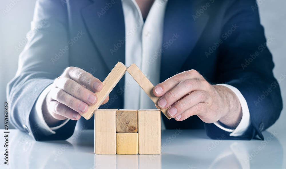 Business man, real estate agent, insurance agent or architect arranging and holding protecting roof over house made from wooden blocks; concept Business man, real estate agent, insurance agent or architect arranging and holding protecting roof over house made from wooden blocks; concept