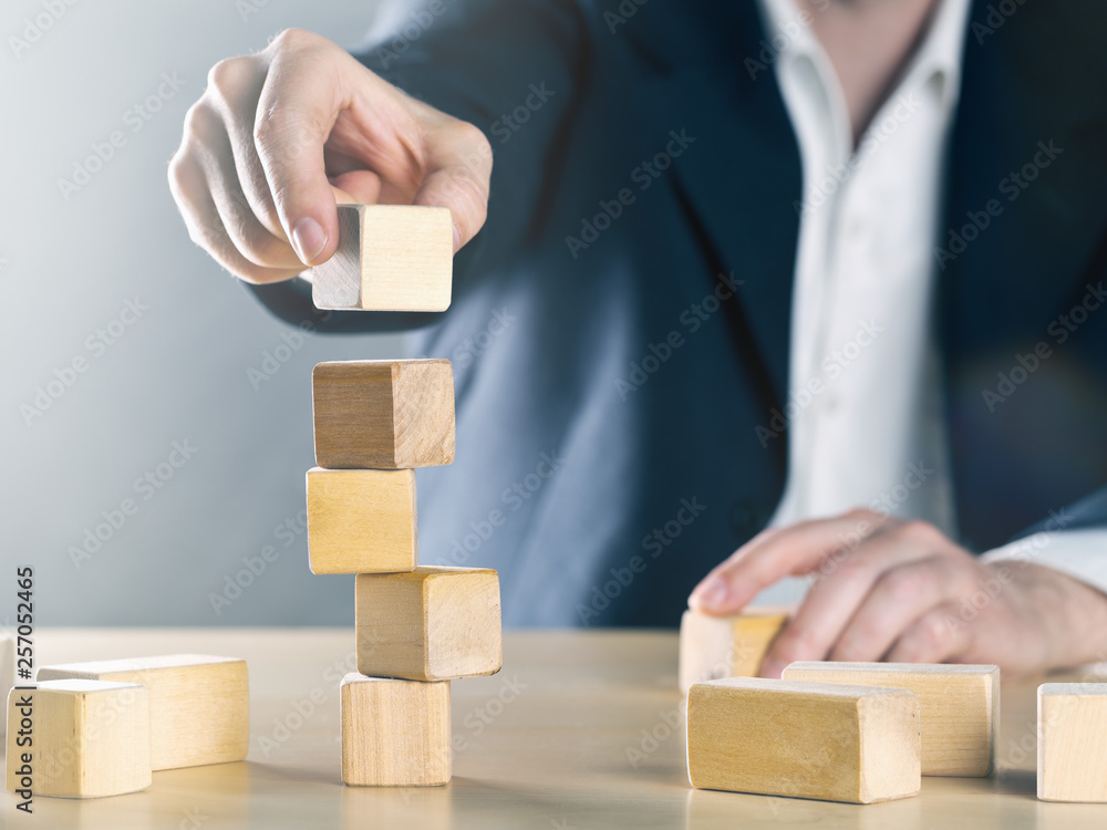 Business man puts next stone on shaky tower structure made from wooden blocks; career or achievement or complex project management concept