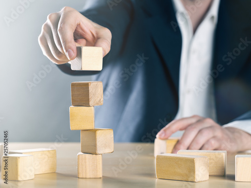 Business man puts next stone on shaky tower structure made from wooden blocks; career or achievement or complex project management concept