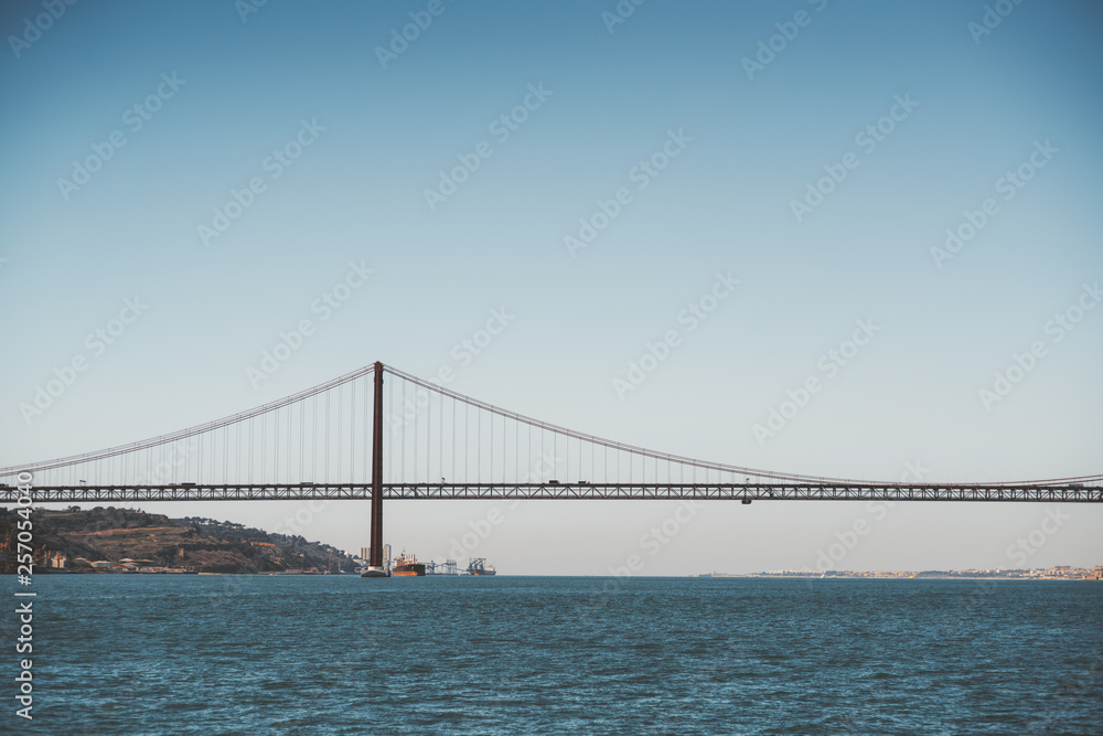 Fototapeta premium Widok z wody ogromnego wiszącego mostu „Ponte 25 de Abril” nad rzeką Tag w Lizbonie, Portugalia w ciepły, poranny dzień z horyzontem w tle