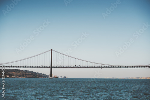 A view from the water of a huge suspension bridge "Ponte 25 de Abril" over the river Tagus in Lisbon, Portugal on a warm morning day with the horizon in the background