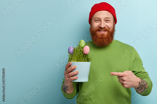 Fotografi Glad foxy hipster male happy to bring colored Easter eggs, points at pot where t