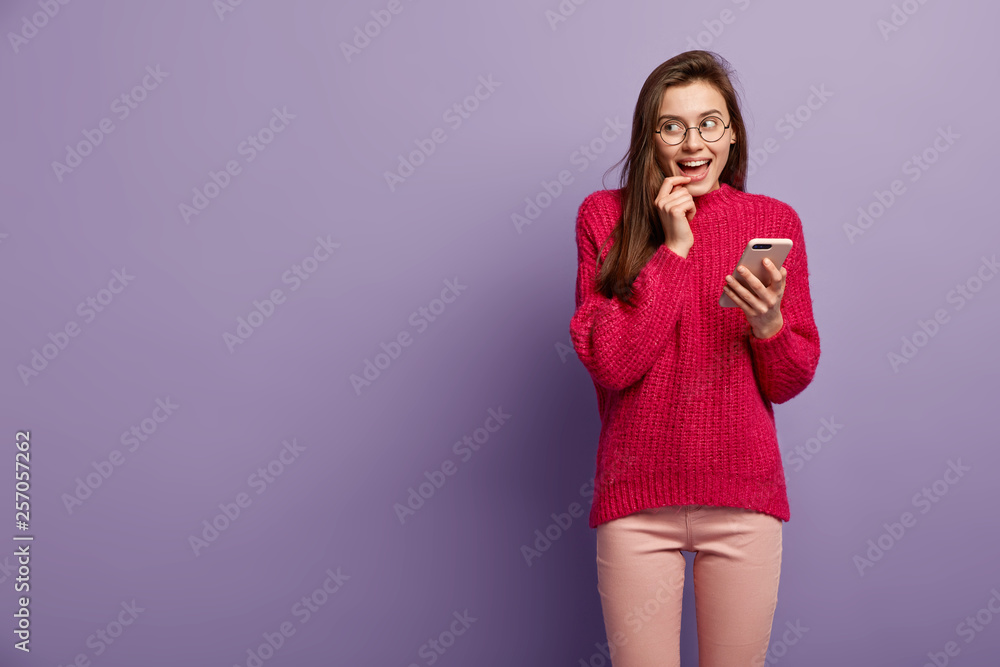 Dreamy cheerful lovely woman uses smart phone for taking pictures, thinks over received notification, looks gladfully aside, surfs internet, wears long knitted sweater, isolated over purple wall Dreamy cheerful lovely woman uses smart phone for taking pictures, thinks over received notification, looks gladfully aside, surfs internet, wears long knitted sweater, isolated over purple wall