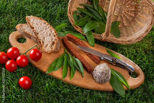 Bärlauch, frisch geerntet auf Holzbrett liegend mit Tomaten, Wurst und Brot, Picknick auf Wiese