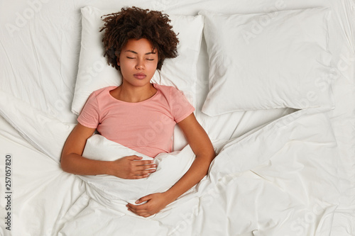Night time concept. Restful dark skinned curly woman has sleep time, slumber and sweet dreams, poses on white fresh linen, rests after tired day, being in resting state, fell into deep sleep