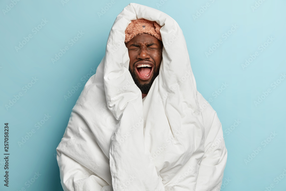 Frustrated sad black man feels stressed to wake up early, wears eyemask on forehead, shouts with negative emotions, wrapped in white soft blanket, isolated over blue background. Bedding concept Frustrated sad black man feels stressed to wake up early, wears eyemask on forehead, shouts with negative emotions, wrapped in white soft blanket, isolated over blue background. Bedding concept
