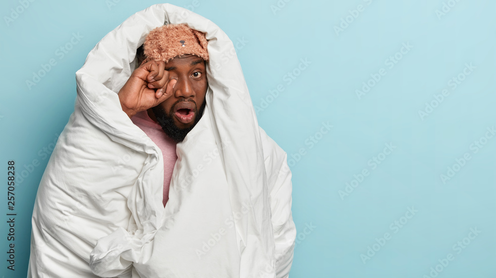 Its difficult to wake up early. Dark skinned sad man rubbs eyes, wants to sleep, wrapped in white blanket, has sleepy tired look, stands over blue wall with free space aside. Awakening concept Its difficult to wake up early. Dark skinned sad man rubbs eyes, wants to sleep, wrapped in white blanket, has sleepy tired look, stands over blue wall with free space aside. Awakening concept
