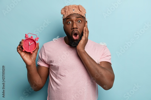 Frightened young black man opens mouth widely, keeps hand on cheek, carries alarm clock, wears eyemask and casual t shirt, isolated over blue background, being late, impressed early in morning photo