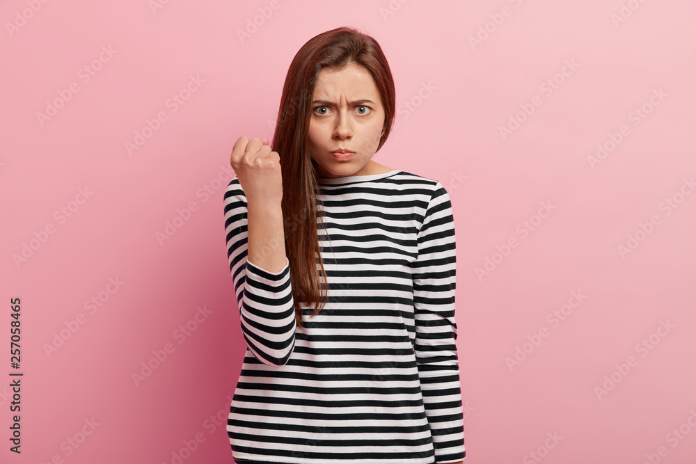 Dont make me annoyed. Furious angry gloomy woman shows fist, has gloomy expression, wears striped sweater, promises to revenge, models over pink background, says I will show you how disobey. Dont make me annoyed. Furious angry gloomy woman shows fist, has gloomy expression, wears striped sweater, promises to revenge, models over pink background, says I will show you how disobey.