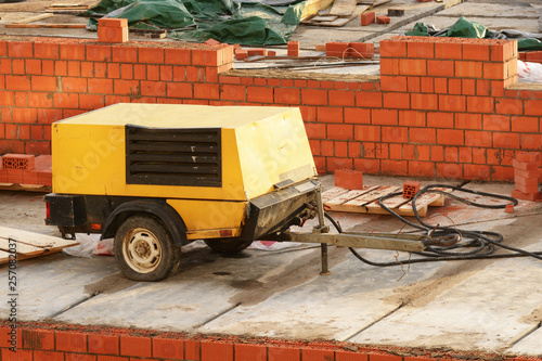 Mobile air compressor on construction site of a red brick house. It's device not new, painted black and yellow. It has pneumatic wheels and a hitch to attach to the car.