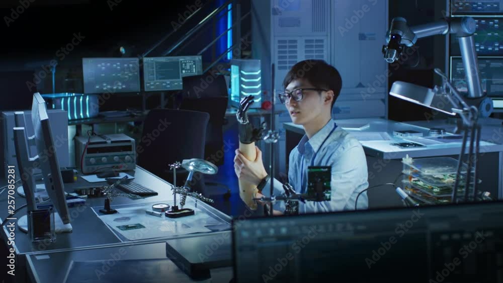 Futuristic Prosthetic Robot Arm Being Tested by a Professional Japanese Development Engineer in a High Tech Research Laboratory with Modern Computer Equipment. Futuristic Prosthetic Robot Arm Being Tested by a Professional Japanese Development Engineer in a High Tech Research Laboratory with Modern Computer Equipment.