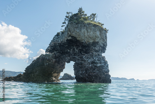 A backlit, offshore, natural arch in a rock stack with trees growing on top, framing another small island. On a sunny, summer's day in Coromandel, New Zealand.