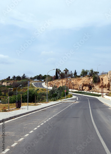 an empty two lame road with pavements and lampposts curving up a hill into the distance with surrounding trees and blue sunlit sky