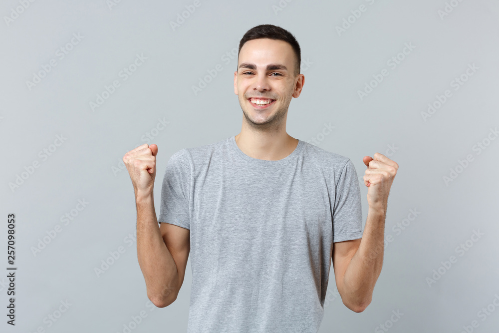 Fototapeta premium Portrait of cheerful young man in casual clothes looking camera, clenching fists like winner isolated on grey wall background in studio. People sincere emotions, lifestyle concept. Mock up copy space.