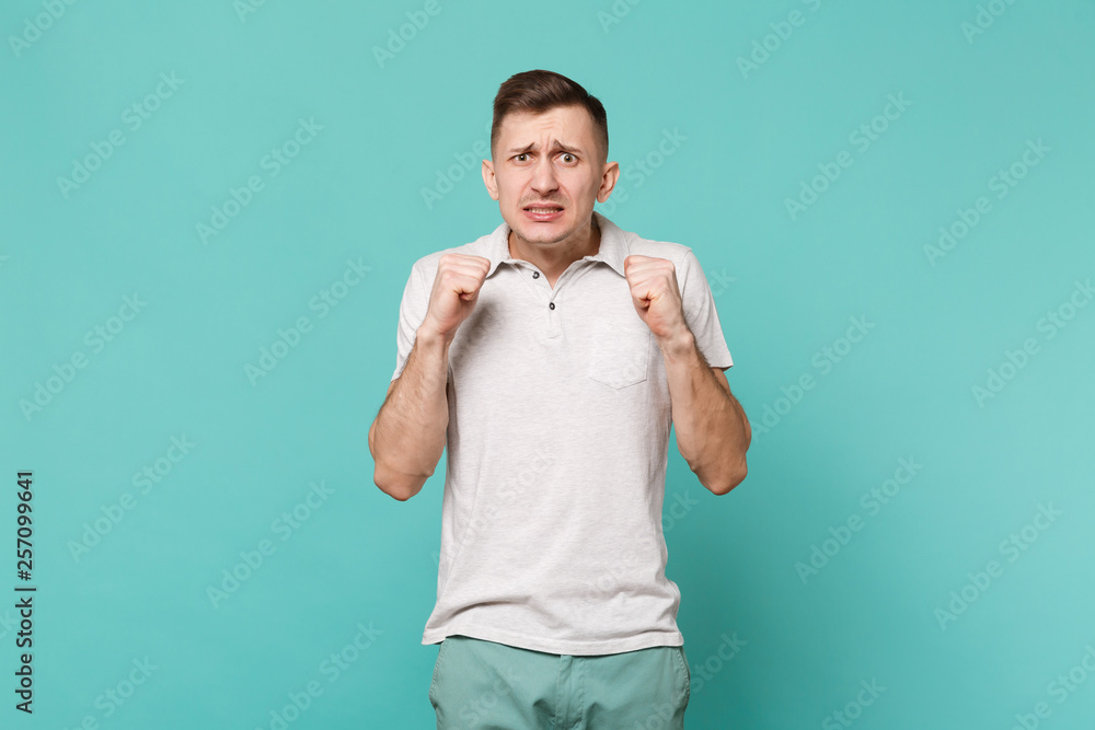 Portrait of waiting young man in casual clothes standing and clenching fists isolated on blue turquoise wall background in studio. People sincere emotions, lifestyle concept. Mock up copy space. Portrait of waiting young man in casual clothes standing and clenching fists isolated on blue turquoise wall background in studio. People sincere emotions, lifestyle concept. Mock up copy space.