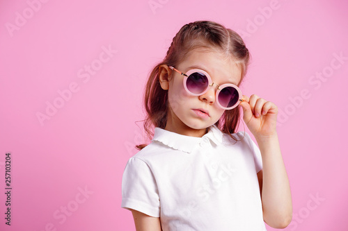 Portrait of beautiful smiling cute model in trendy round sunglasses. Casual 7 y.o. girl posing isolated on pink background.