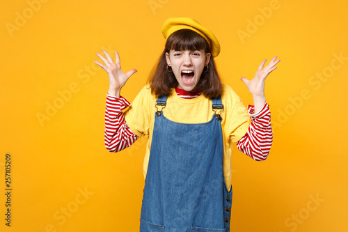 Portrait of crazy screaming girl teenager in french beret, denim sundress spreading hands isolated on yellow wall background in studio. People sincere emotions, lifestyle concept. Mock up copy space.