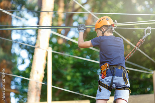 Sporty, young, cute boy in white t shirt spends his time in adventure rope park in helmet and safe equipment in the park in the summer. Active lifestyle concept, summer camp
