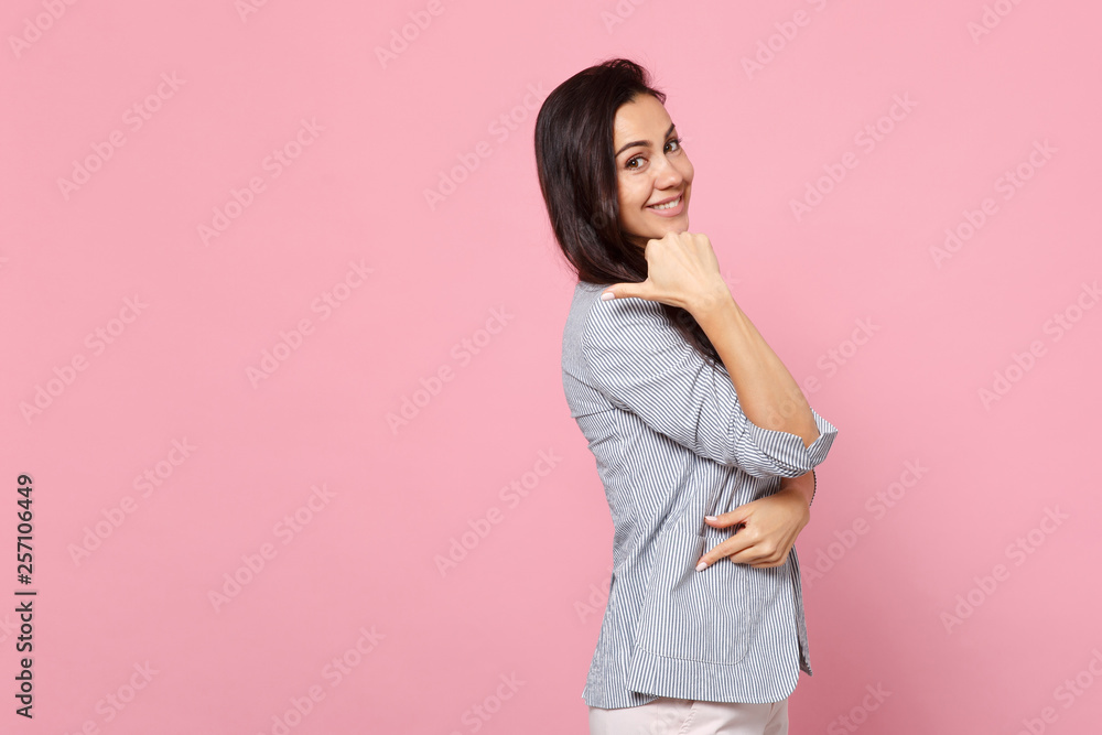 Side view of smiling attractive young woman in striped jacket pointing thumb aside isolated on pink pastel wall background in studio. People sincere emotions, lifestyle concept. Mock up copy space. Side view of smiling attractive young woman in striped jacket pointing thumb aside isolated on pink pastel wall background in studio. People sincere emotions, lifestyle concept. Mock up copy space.