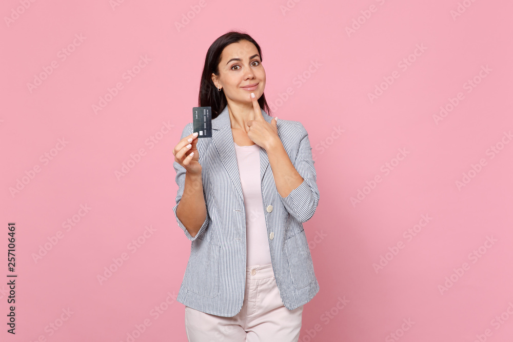 Smiling young woman in striped jacket putting finger on chin holding credit bank card isolated on pink pastel wall background in studio. People sincere emotions, lifestyle concept. Mock up copy space. Smiling young woman in striped jacket putting finger on chin holding credit bank card isolated on pink pastel wall background in studio. People sincere emotions, lifestyle concept. Mock up copy space.
