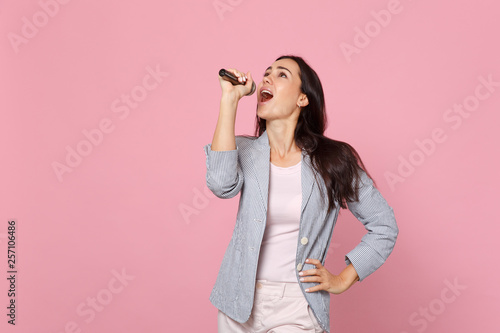 Portrait of attractive young woman in striped jacket looking up, sing song in microphone isolated on pink pastel background in studio. People sincere emotions, lifestyle concept. Mock up copy space.