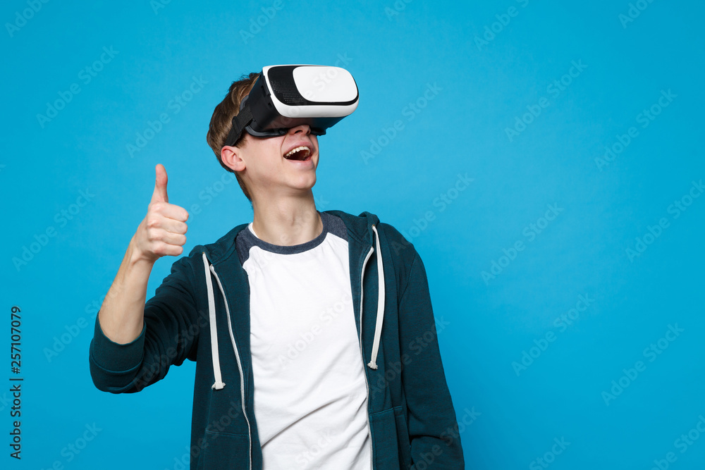 Cheerful young man in casual clothes looking on headset showing thumb up isolated on blue wall background in studio. People sincere emotions, lifestyle concept. Mock up copy space. Advertising area. Cheerful young man in casual clothes looking on headset showing thumb up isolated on blue wall background in studio. People sincere emotions, lifestyle concept. Mock up copy space. Advertising area.