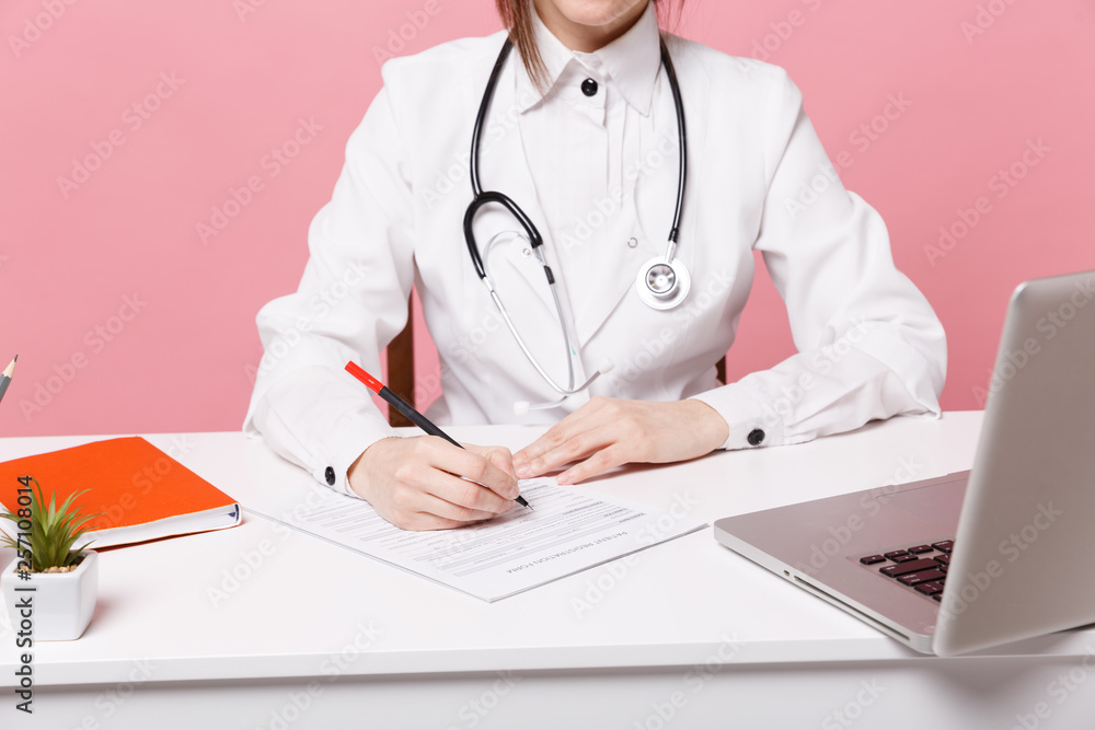 Cropped female doctor siting at desk working filling out medical documents in hospital isolated on pastel pink wall background indoors. Woman in medical gown stethoscope. Healthcare medicine concept. Cropped female doctor siting at desk working filling out medical documents in hospital isolated on pastel pink wall background indoors. Woman in medical gown stethoscope. Healthcare medicine concept.