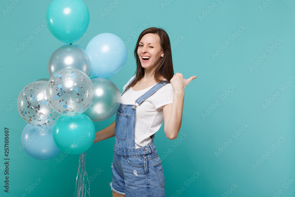 Fototapeta premium Cheerful young woman in denim clothes blinking, pointing thumb aside celebrating hold colorful air balloons isolated on blue turquoise wall background. Birthday holiday party, people emotions concept.