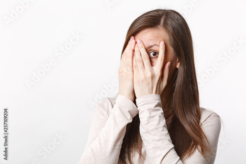 Portrait of peeping pretty young woman in light clothes hiding, covering face eith hands isolated on white wall background in studio. People sincere emotions, lifestyle concept. Mock up copy space.