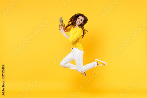 Stunning young woman in casual clothes jumping, hold fresh ripe pineapple fruit isolated on yellow orange wall background in studio. People vivid lifestyle, relax vacation concept. Mock up copy space.
