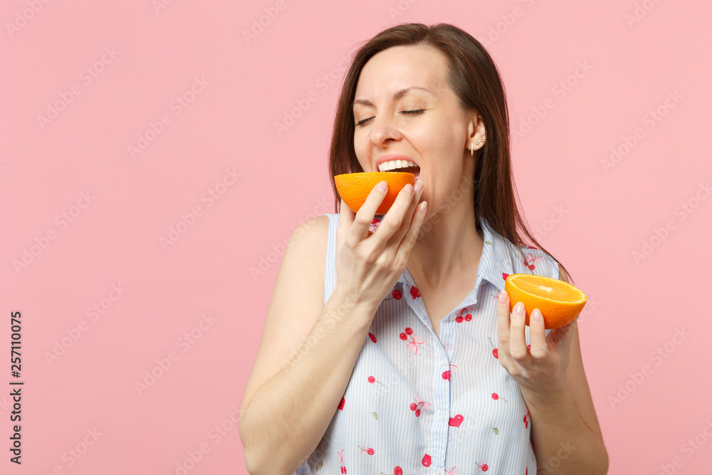 Pretty young woman in summer clothes keeping eyes closed biting half of fresh ripe orange fruit isolated on pink pastel background. People vivid lifestyle relax vacation concept. Mock up copy space.