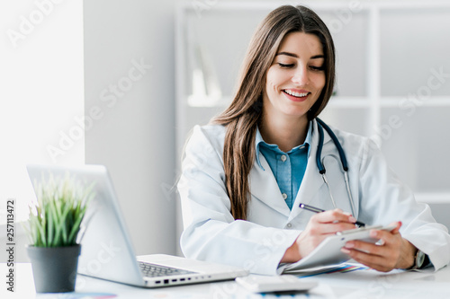 Photography Smiling young female doctor working at the clinic , she is using a computer and writing medical reports