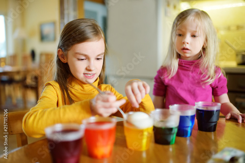 Two cute young sisters dyeing Easter eggs at home. Children painting colorful eggs for Easter hunt. Kids getting ready for Easter celebration.