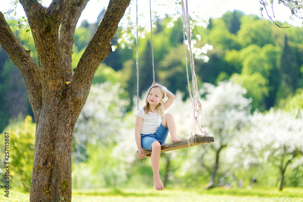 Cute little girl having fun on a swing in blossoming old apple tree garden outdoors on sunny spring day.