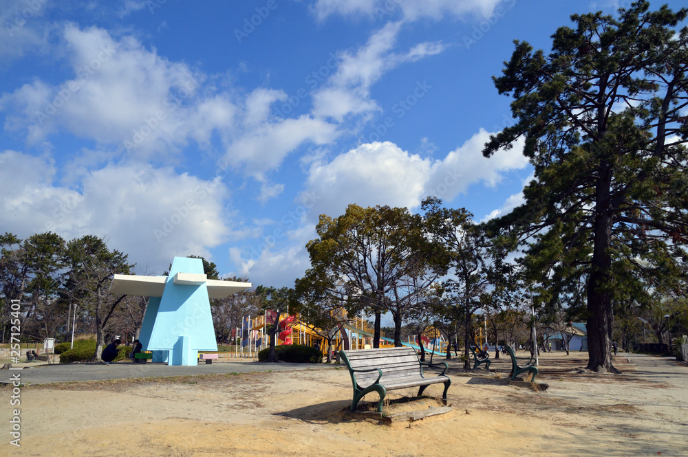 白い雲が浮かぶ青空と、子どもたちが遊ぶ遊具を背景に、陽射しをあびたベンチがある公園の風景