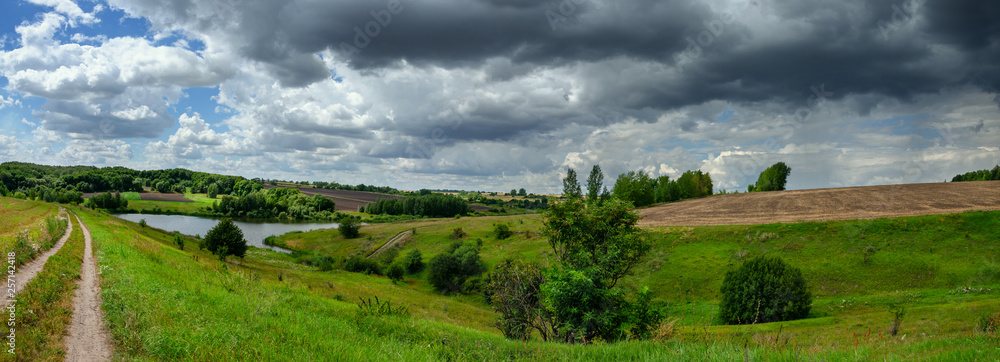 Plakat Cloudy windy summer panoramic landscape with ground country road and green hills.
