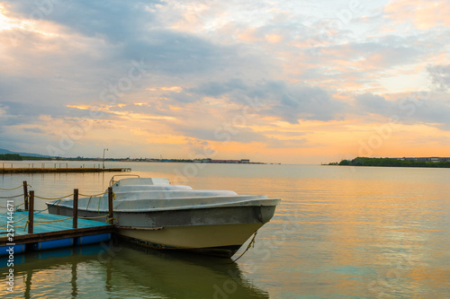 Old white fishing boat docked by the plank on a lagoon and pastel colors illuminate the sky at sunset.