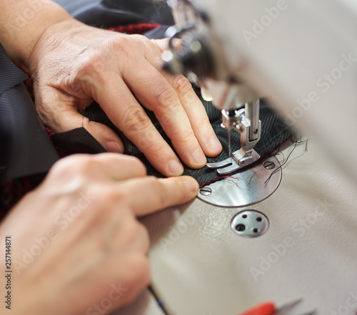 Top view of woman hands stitching garment on professional sewing machine at workplace. Tailor hands holding chequered textile for skirt production. Blurred background. Close up view of sewing process.