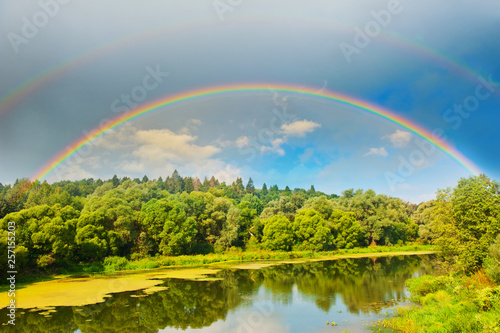 Bright double rainbow in the sky with clouds above the forest and the river