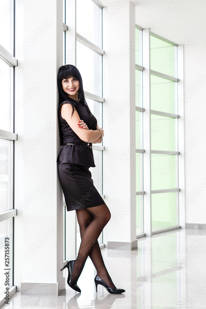 Young Beautiful happy brunette woman dressed in a black business suit with a short skirt standing near the window in office, smiling, looking to camera. Young Beautiful happy brunette woman dressed in a black business suit with a short skirt standing near the window in office, smiling, looking to camera.